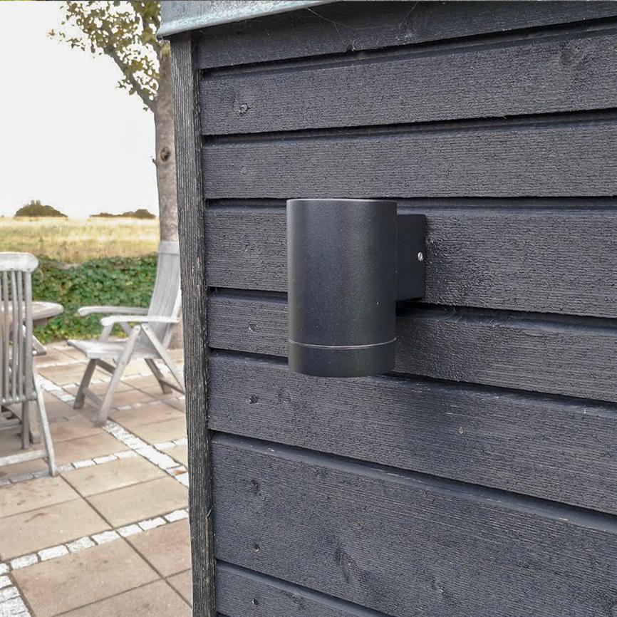 En cylindrisk sort væglampe, slukket, er monteret på mørk, vandret træbeklædning; i baggrunden ses en flisebelagt terrasse med udendørsstole og et åbent græsareal.