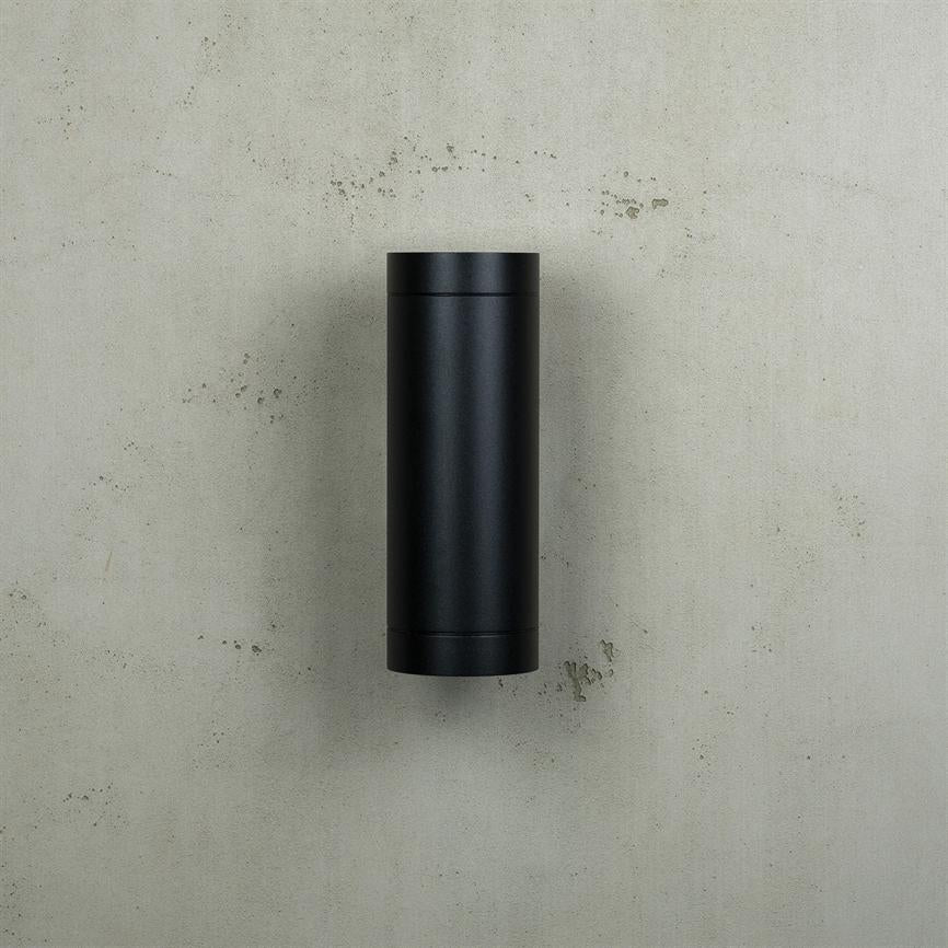 Sort cylindrisk beholder er centreret og monteret på en lysegrå, let plettet betonvæg med små afskalninger.