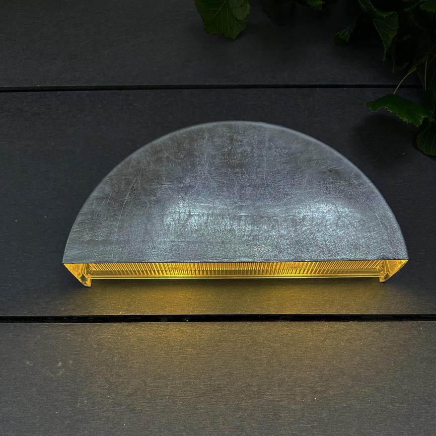 Halvcirkelformet metallampe udsender varmt, gult lys nedad og kaster en stribe på mørke vandrette paneler; grønne blade ses i øverste højre hjørne.