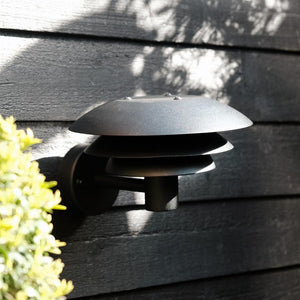 En sort, lagdelt væglampe er monteret på en mørk trævæg og står slukket; sollys kaster skygger af løv, og grønne blade ses i venstre forgrund.