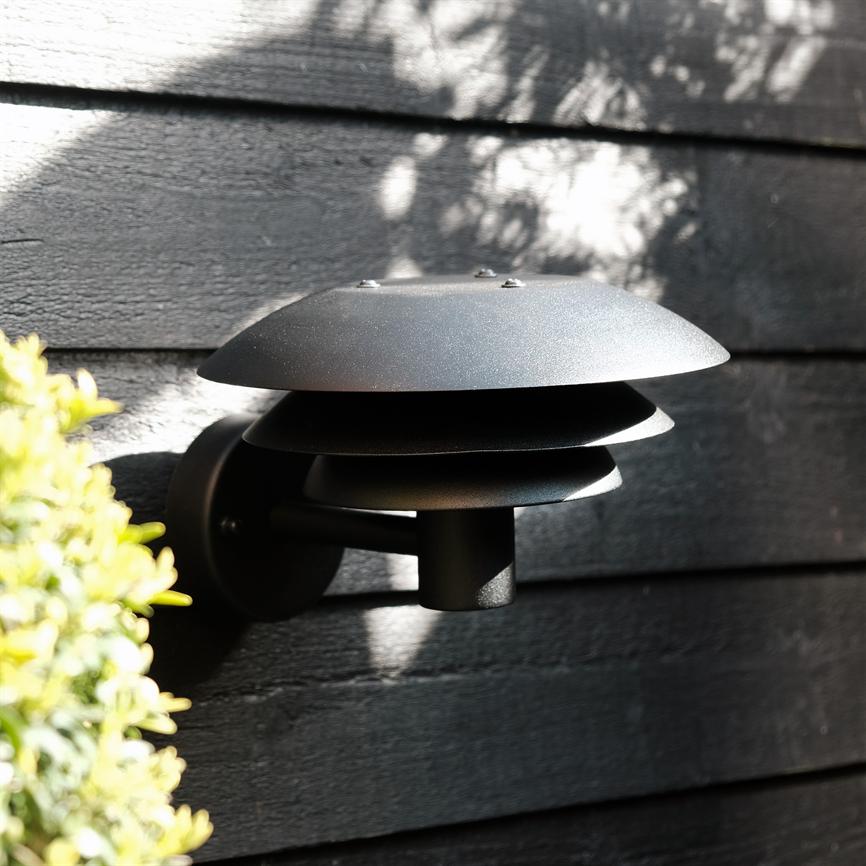 En sort, lagdelt væglampe er monteret på en mørk trævæg og står slukket; sollys kaster skygger af løv, og grønne blade ses i venstre forgrund.