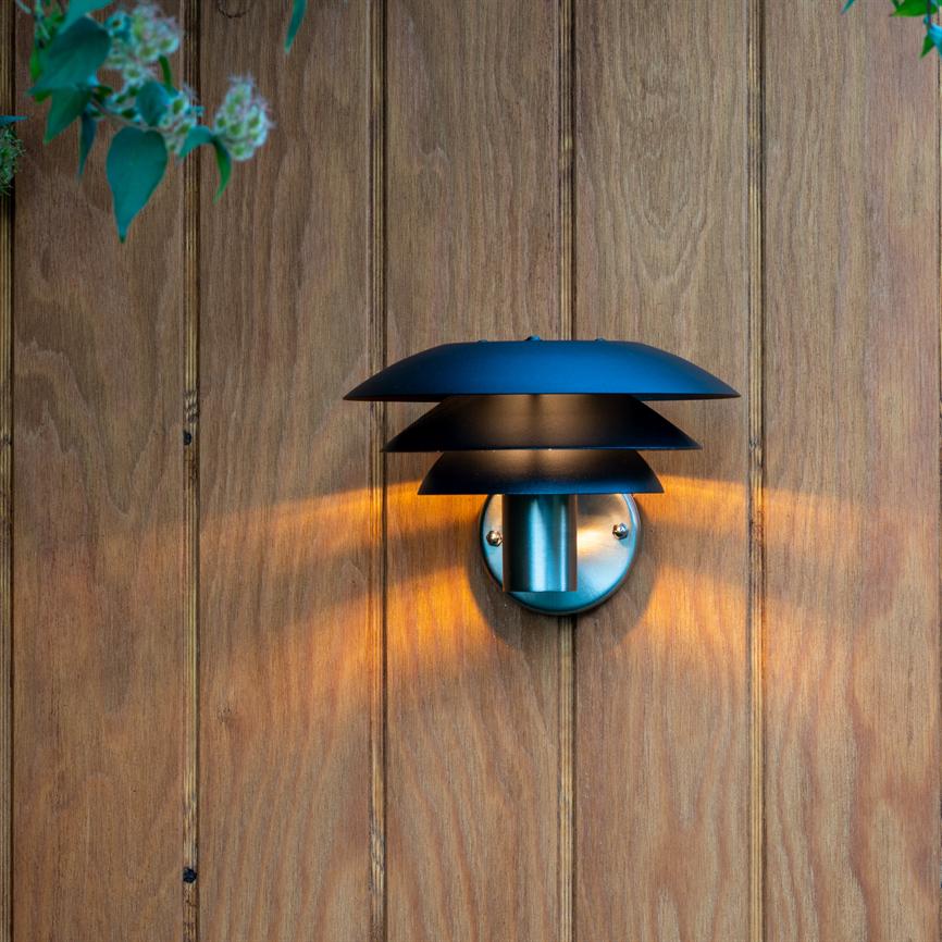 Sort, skiveopbygget væglampe udsender varmt, diffust lys gennem tre skærme, monteret på en lodret, lyst træbeklædt væg; grønne blade ses i øverste hjørner.