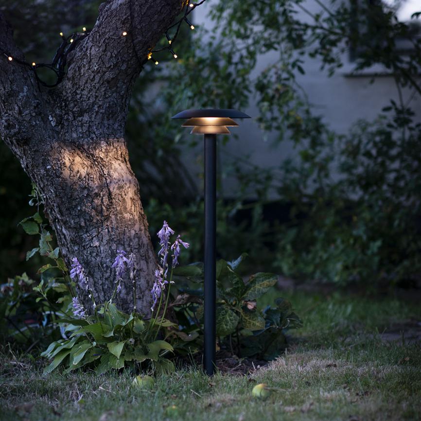 En sort havelygte står tændt og kaster varmt, diffust lys over bunddække og lilla blomster ved en tyk træstamme, omgivet af grønt løv, græsplæne og diskrete lyskæder.