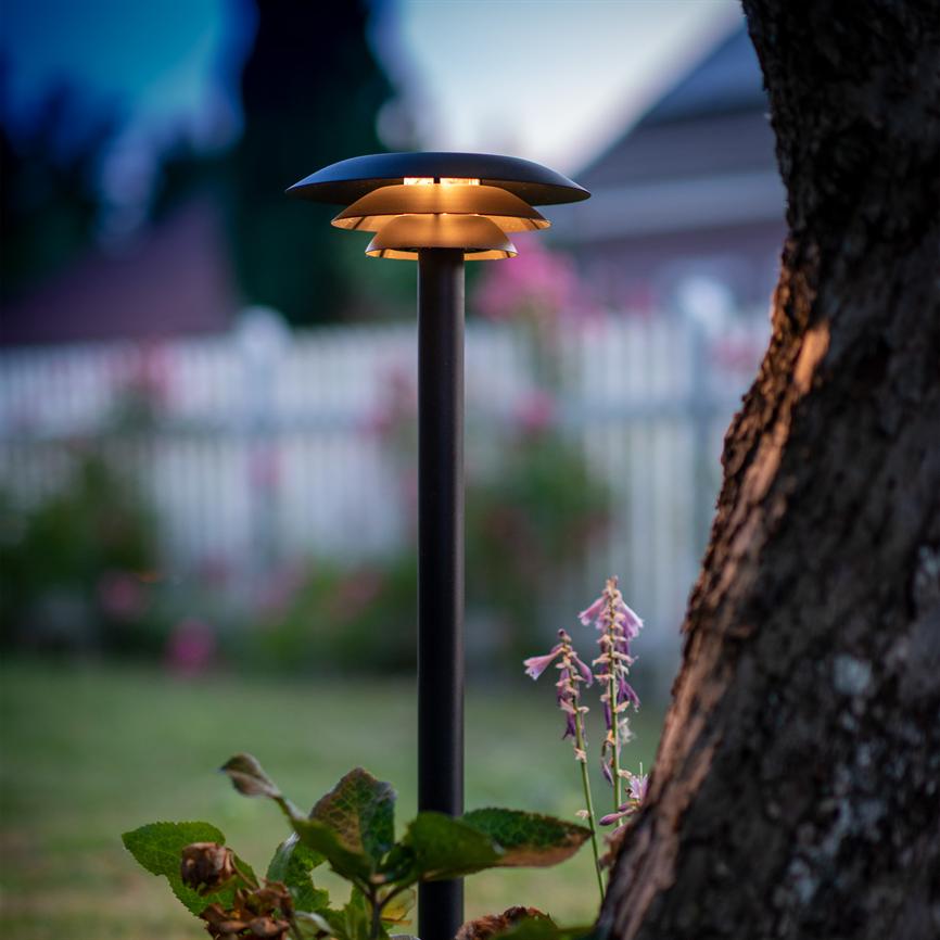 En sort havebelysningslampe sender varmt, nedadrettet lys, stående ved en træstamme og blomster i forhaven. Baggrund: sløret hvidt stakit og aftenskyer ved tusmørke.