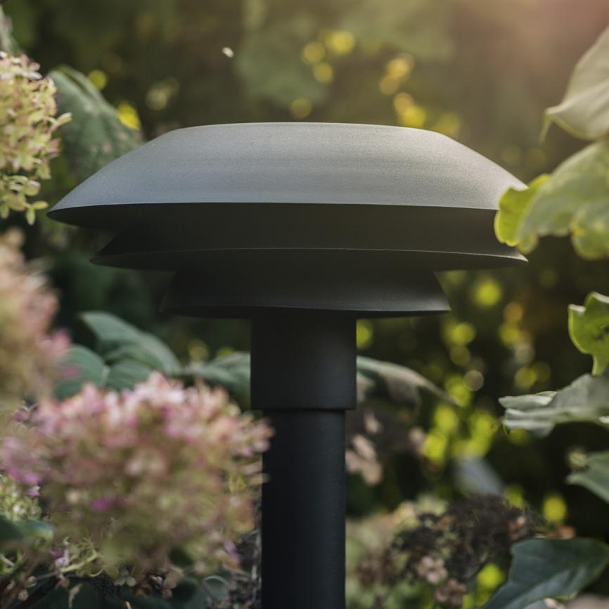 En sort, svampelignende havebelysningslampe står på en tyk søjle, placeret blandt grønne blade og lyserød-hvide blomster i en soloplyst havebaggrund.