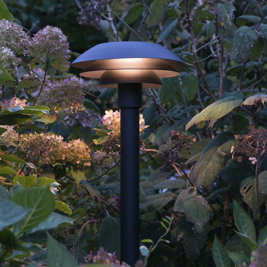 En sort, svampelignende havelygte kaster varmt, blødt skær nedad blandt frodige blade og visne hortensiaer i en skumringshave.