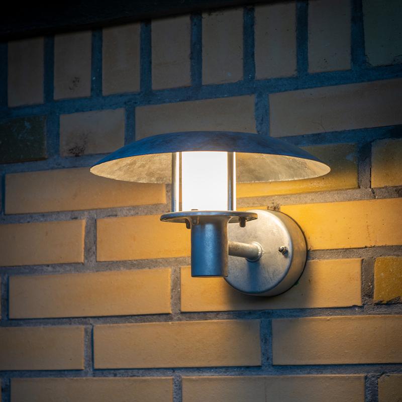 Rund metal væglampe udstråler varmt, hvidt lys gennem en cylindrisk glasskærm og oplyser en gulbrun teglstensvæg uden synlig tekst.