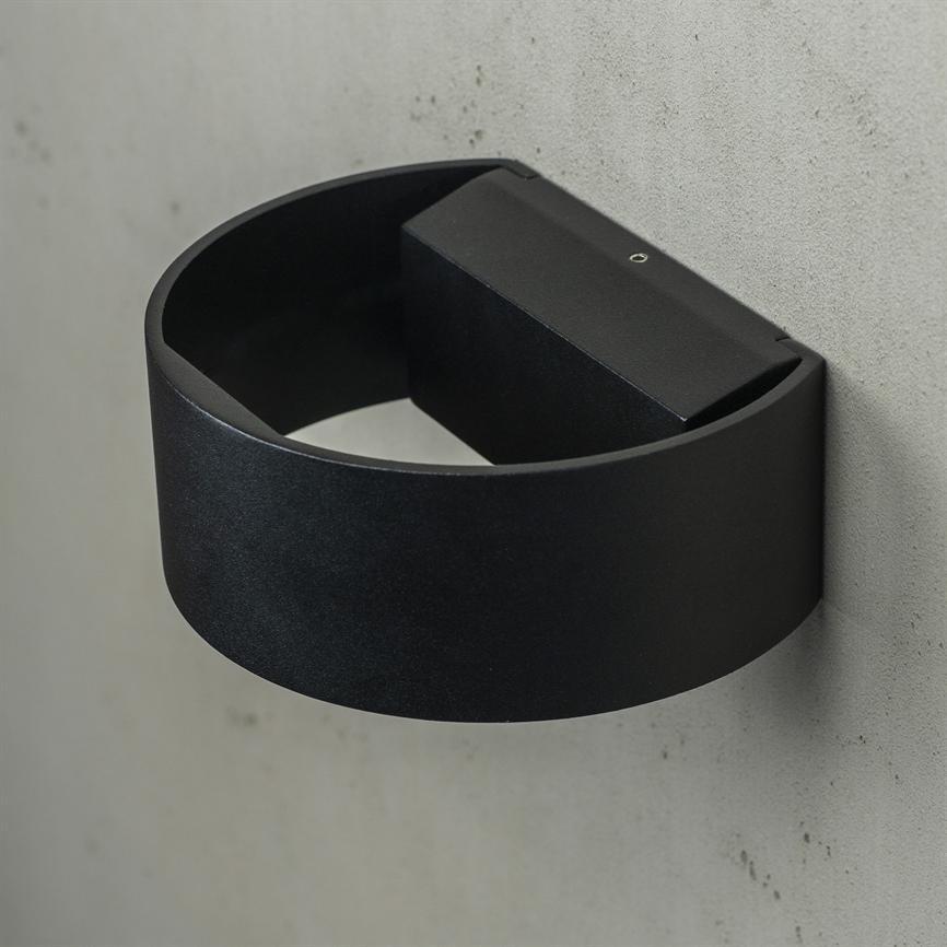 Sort, halvcirkelformet væglampe sidder monteret på en grå betonvæg, åben nedad og vinklet ind mod væggen; synlig rektangulær monteringsboks bagpå.