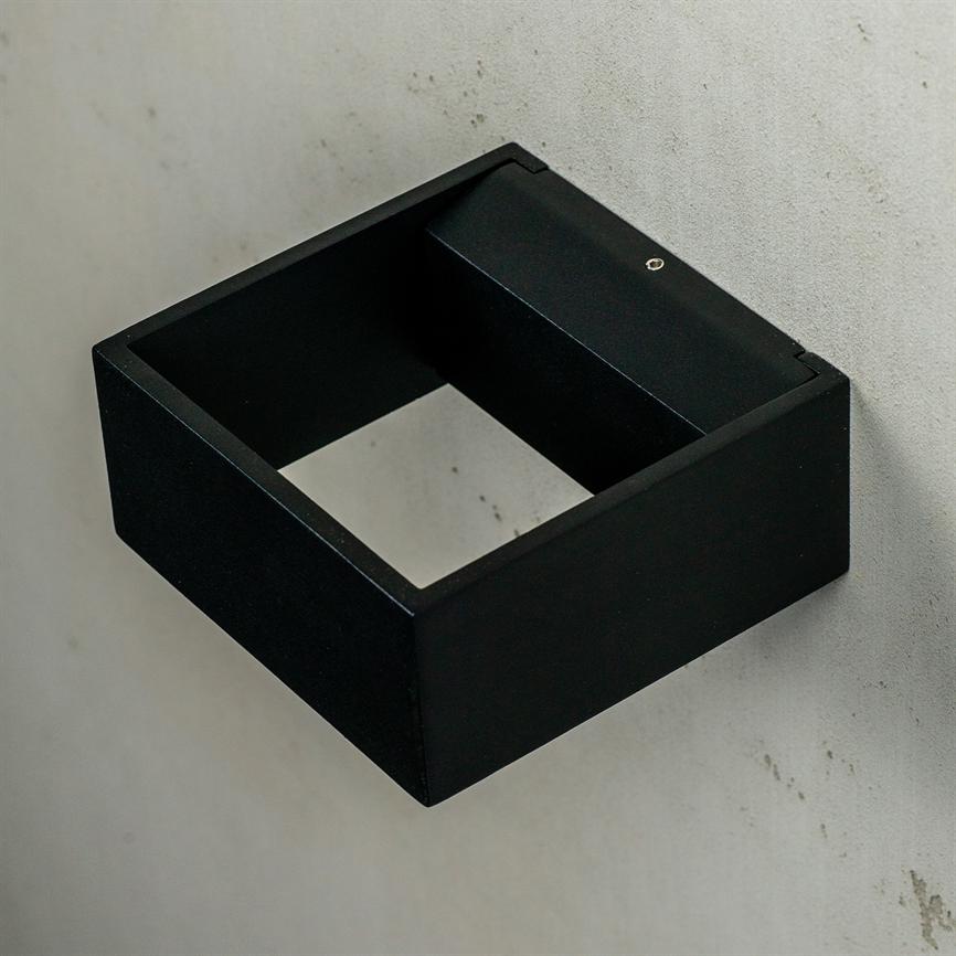 Sort, firkantet metalramme med åben midte; den er monteret på en lysgrå, let plettet væg med en synlig skrue, og rammen fremstår tom og mat.