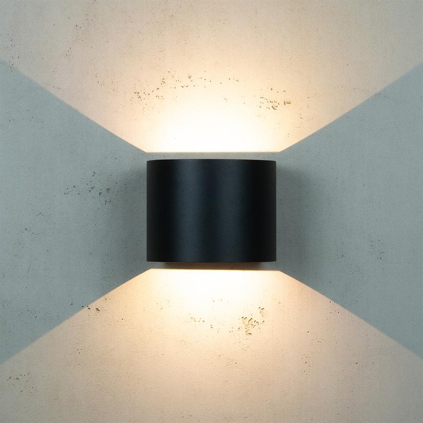 Cylindrisk sort væglampe udsender varmt lys opad og nedad, kaster krydsformede lystriangler på en ru, lysegrå betonvæg.