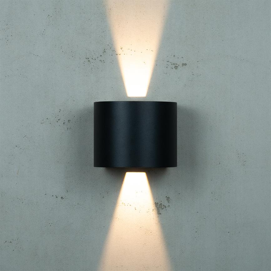 Sort cylindrisk væglampe udsender to koniske lysstråler opad og nedad, der kaster varmt, diffust lys mod en ru, lysegrå betonvæg med små pletter og ujævnheder.