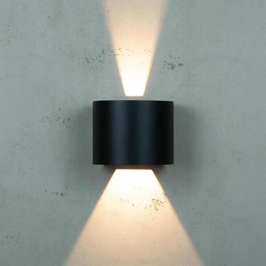 En sort cylindrisk væglampe kaster symmetriske, varme lysstråler opad og nedad på en rå, lysegrå betonvæg.