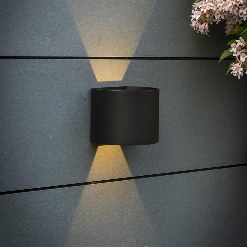 En sort cylinderformet væglampe udsender varme, kegleformede lysstråler opad og nedad; monteret på mørkegrå, horisontalt panelerede væg, med lyserøde blomster i øverste højre hjørne.