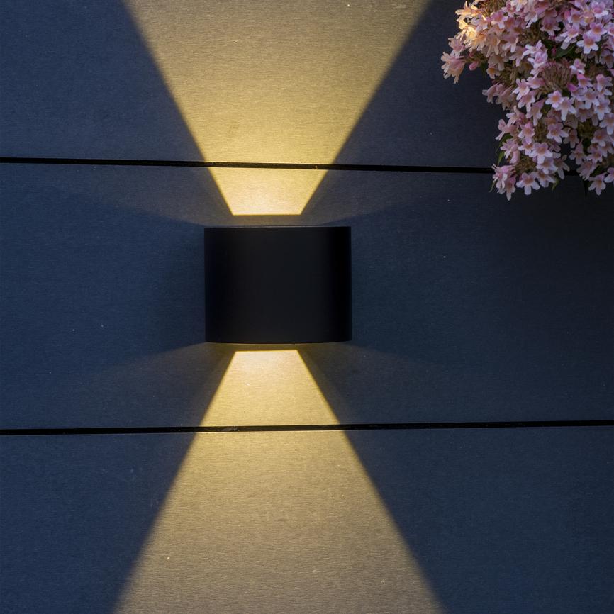 Sort cylindrisk væglampe kaster symmetriske, varme lysstråler op og ned mod mørke, horisontale paneler, med en klynge lyserøde blomster i øverste højre hjørne.