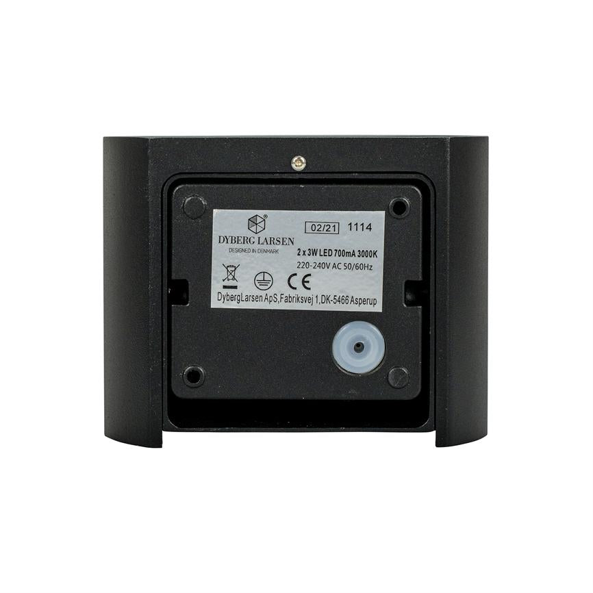 Et rektangulært mærkat (objekt) angiver tekniske data og serienummer (aktion) på bagsiden af en sort væglampe, placeret centralt mod en hvid baggrund (kontekst).
 
 Transkription af teksten:
 DYBERG LARSEN
 DESIGNED IN DENMARK
 
 02/21 1114
 
 2 x 3W LED 700mA 3000K
 220-240V AC 50/60Hz
 
 DybergLarsen ApS, Fabriksvej 1, DK-5466 Asperup