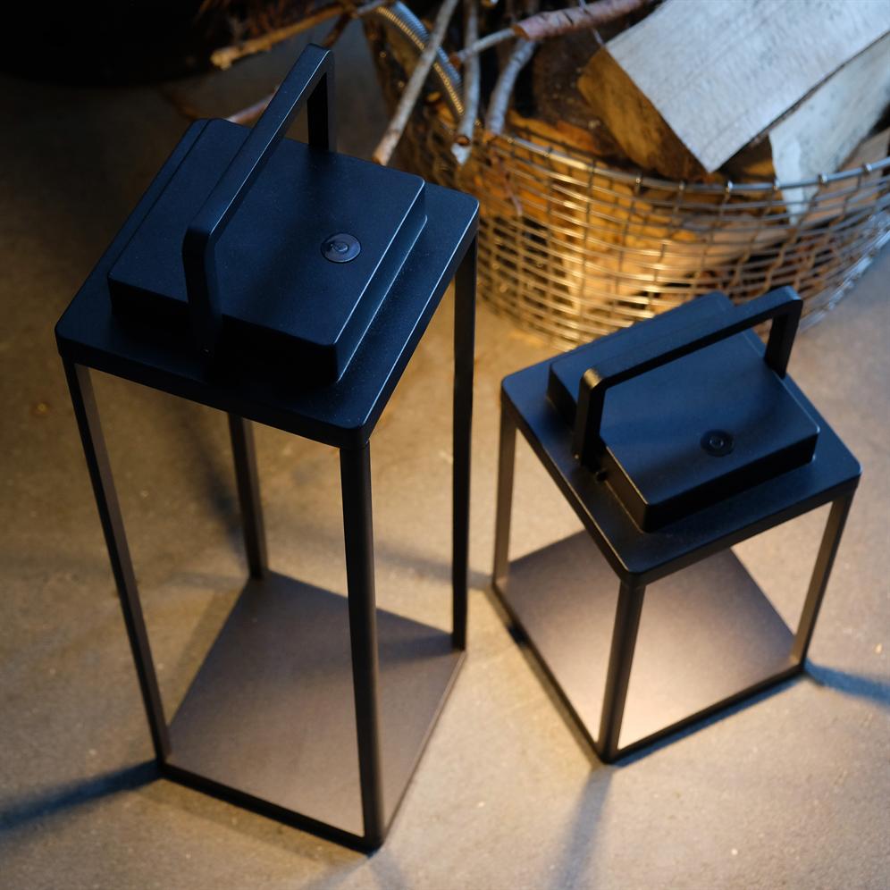 To sorte metal-lanterner står på et betongulv og kaster svage skygger; bag dem står en trådkurv fyldt med brænde.