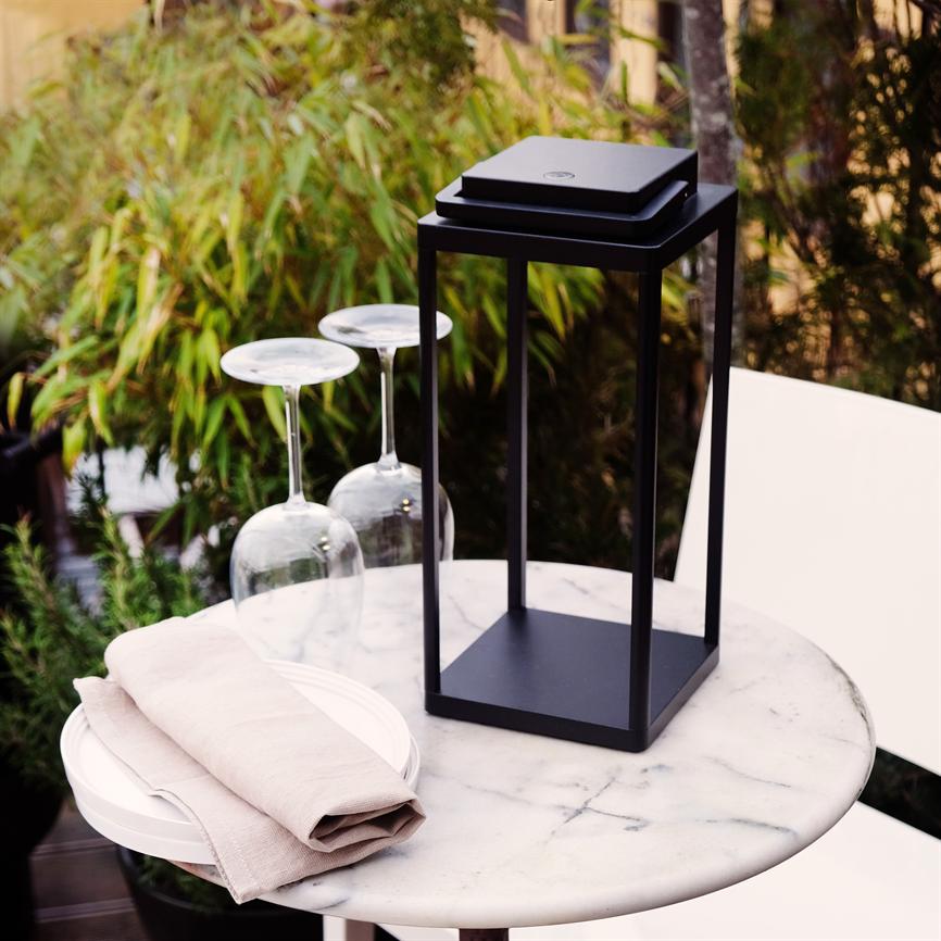 Sort metal-lanterne står på et rundt marmorbord på en udendørs terrasse, flankeret af to opadvendte vinglas, en sammenfoldet beige stofserviet og grønne planter i baggrunden.
