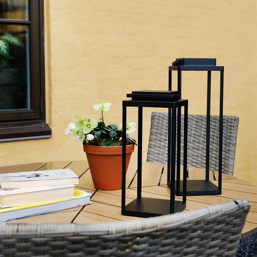 To sorte, firkantede metallanterner står på et træbord; en terrakottakrukke med hvide blomster og en stak bøger ligger ved siden af, flettede stole og en gul murstensvæg i baggrunden.