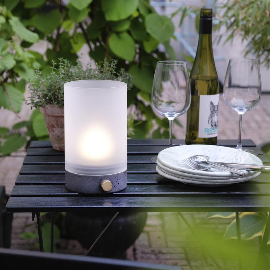 Cylindrisk bordlampe udstråler blødt lys; står på sort træbord ved to vinglas, en vinflaske og en stak tallerkener med bestik, omgivet af grønne planter. Tekst på etiketten: "RIES".