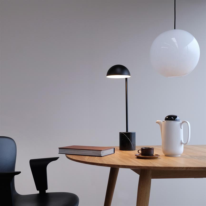 Et rundt træbord står med en brun bog, en sort bordlampe, en kop på en underkop og en hvid termokande; en sort stol ved siden af under en hvid pendel.