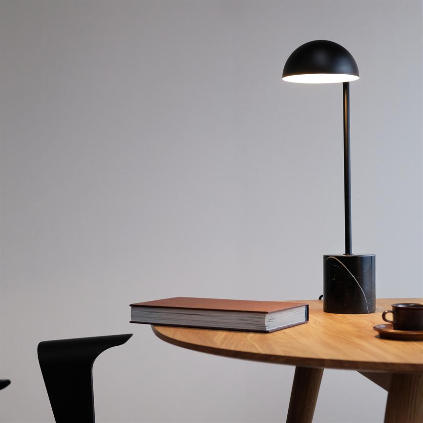 En sort bordlampe lyser over et rundt træbord med en lukket bog og en kaffekop; en sort stol står ved bordkanten i et neutralt, lysegråt rum.