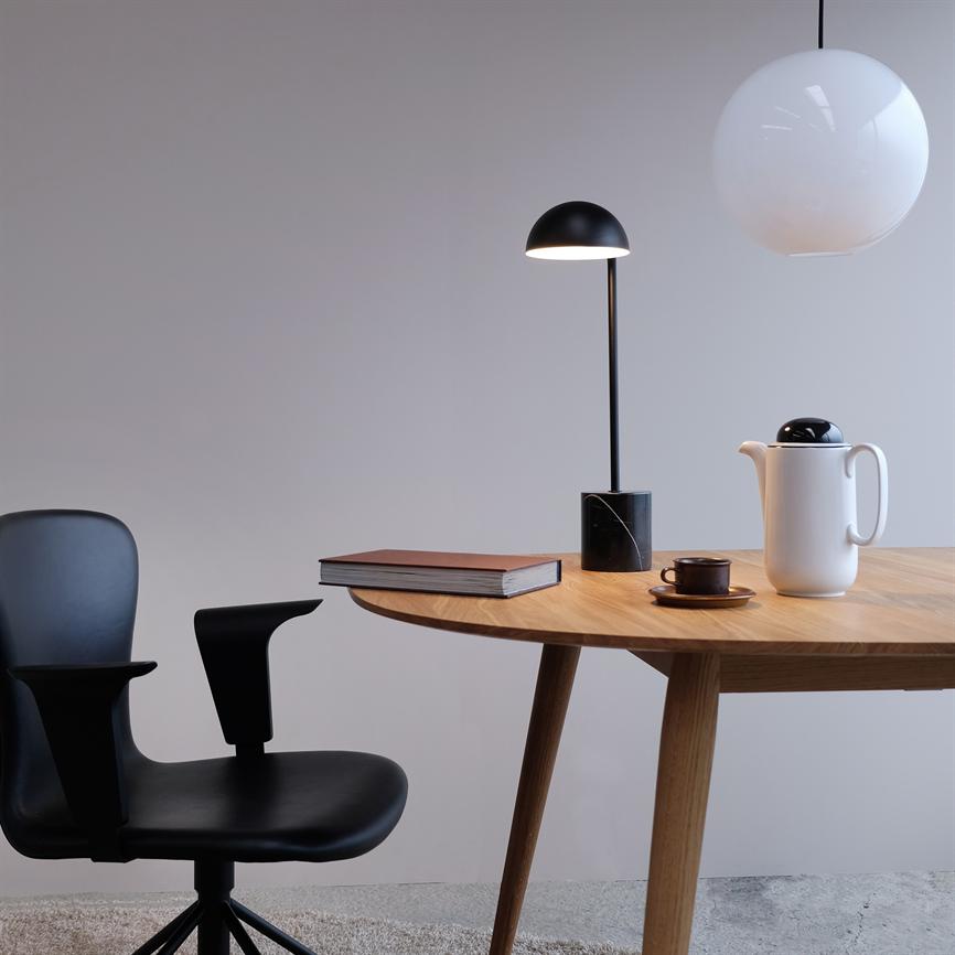 Rundt træbord rummer notesbog, sort bordlampe, hvid termokande og kaffekop; et sort kontorstol står til venstre, hænger en hvid kuglelampe fra loftet, neutral lysegrå baggrund.