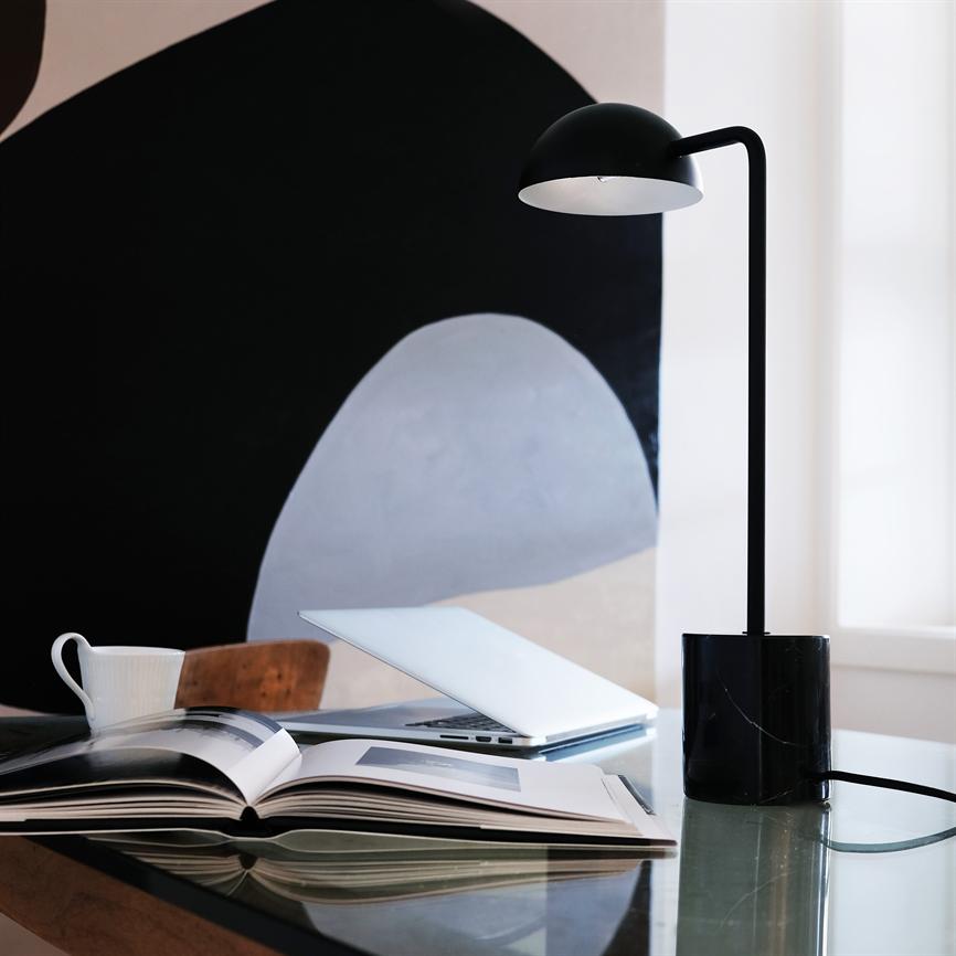 Skrivebordslampe står tændt over et glasbord, kaster lys på en åben bog, en halvåben bærbar og en kaffekop i et moderne, minimalistisk arbejdsrum med abstrakt vægmaleri.