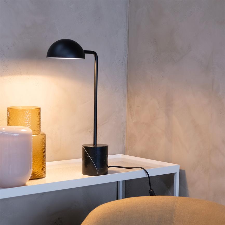 Sort bordlampe lyser varmt nedad og kaster skygge på en tekstureret beige væg; den står på en hvid hylde med to vaser (gul og lyserød) ved en beige stol.