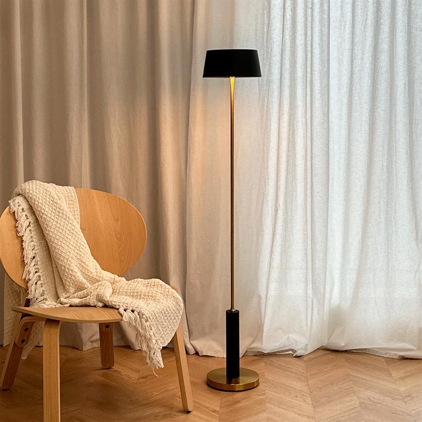 En slank gulvlampe står tændt og kaster varm glød; ved siden af en enkel træstol med et cremefarvet, flettet tæppe; lyse gardiner og parketgulv i baggrunden.