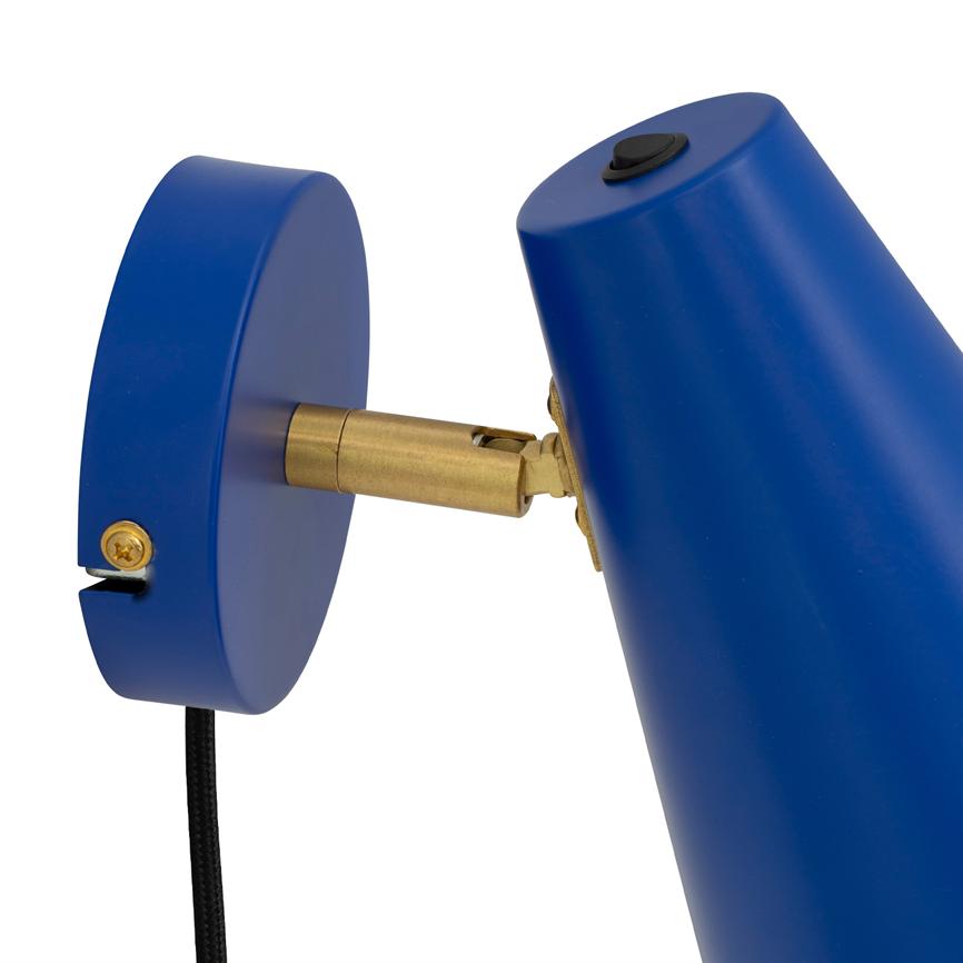 Blå væglampe er vinklet via et messingled og fastgjort til en rund vægplade; en sort stofledning hænger nedad mod en hvid baggrund.