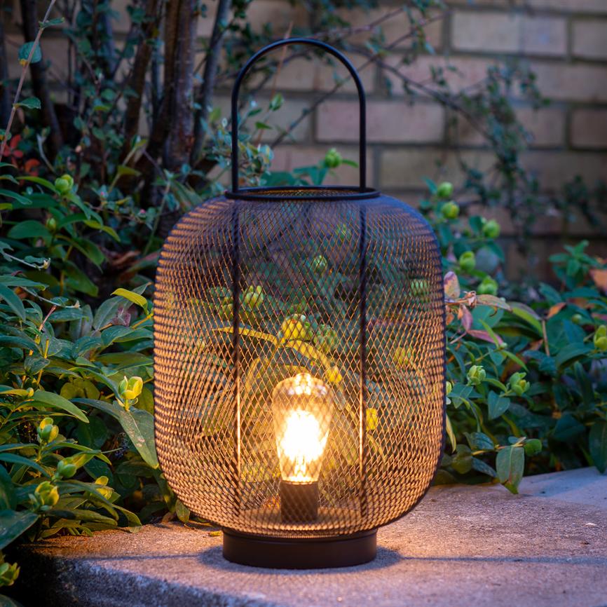 En sort metalgitterlanterne udsender varmt, blødt lys fra en tændt pære; den står på en betonflise omgivet af lave grønne planter foran en murstensvæg.
