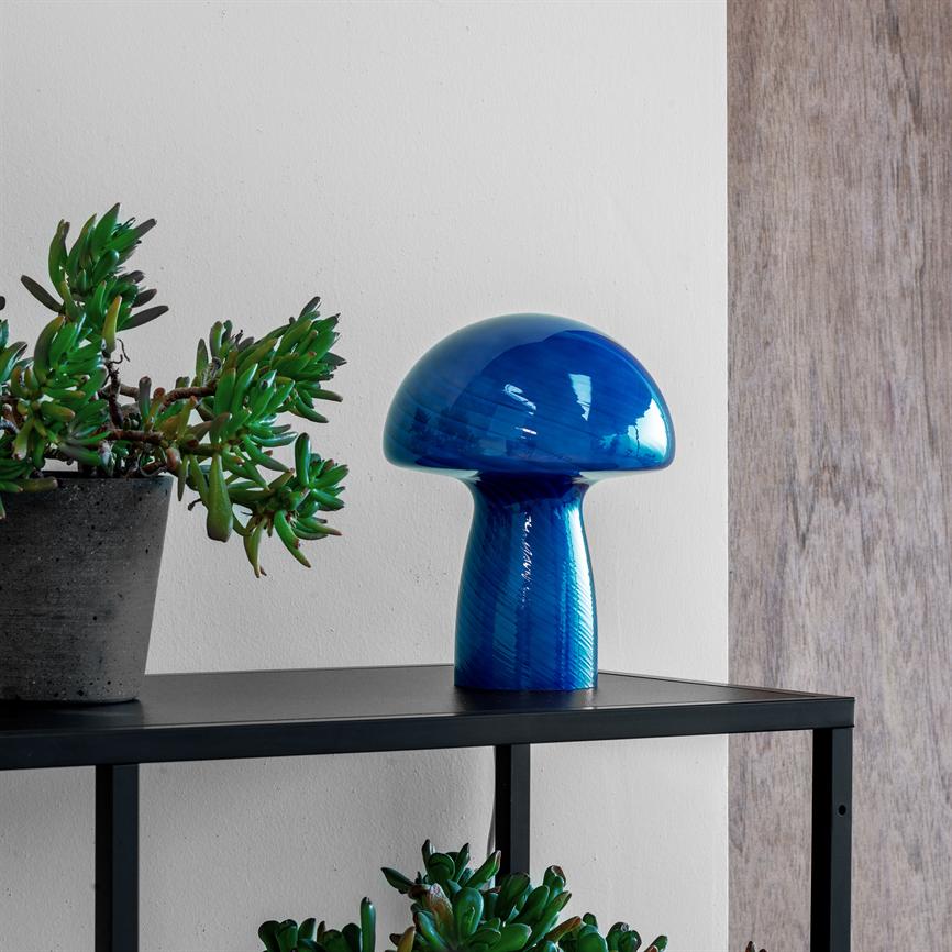 Blå, svampeformet glasobjekt står på en sort metalhylde ved siden af en grå potte med sukkulenter; hvid væg bagved og lyst træpanel til højre.