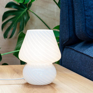 En hvid, spiralmønstret bordlampe lyser blødt på et lyst træbord ved en blå sofa, med grønne stueplanteblade i baggrunden.