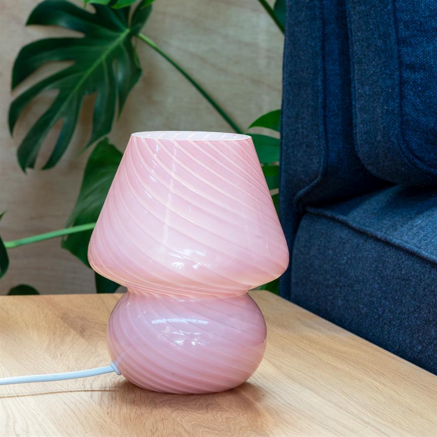 Rosa glasbordlampe med snoede riller står slukket på et lyst træbord, ved siden af en mørkeblå sofa og en grøn stueplante.