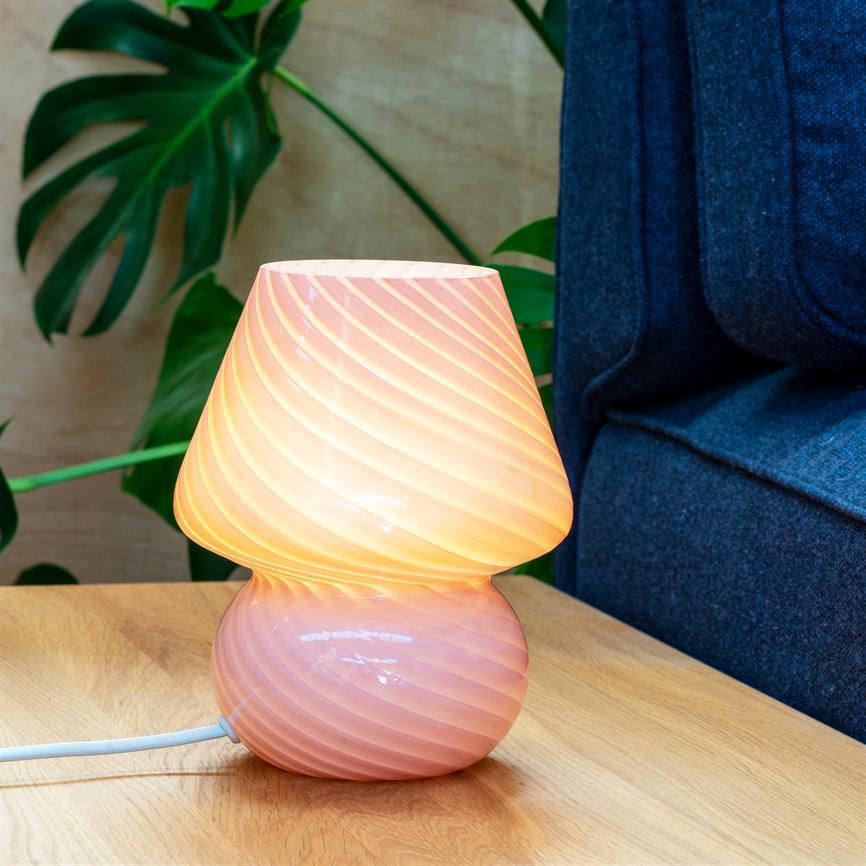 Lampe i rosa glas lyser med en varm, spiralformet glød; den står på et lyst træbord ved en mørkeblå sofa, med store grønne Monstera-blade i baggrunden.