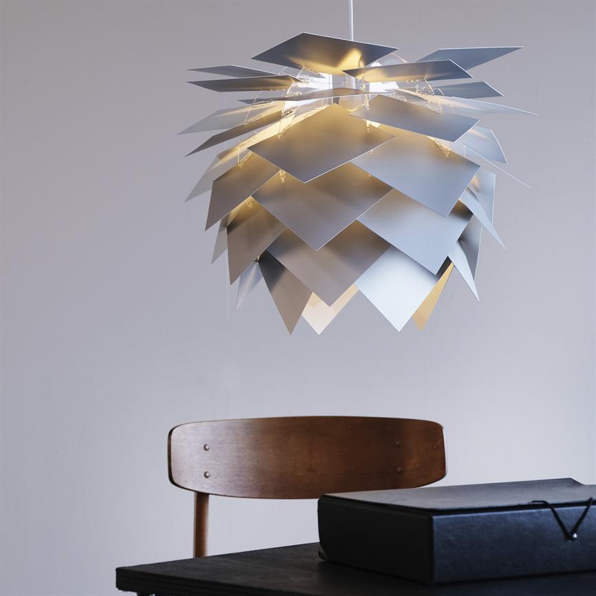 En moderne pendelarmatur formet af overlappende kvadratiske metalplader, udsender varmt lys, hænger over et mørkt bord med en træstol og en sort æske i et neutralt rum.