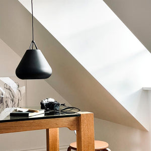 En sort lampe hænger over et træbord; et kamera og nogle bøger ligger på glasbordpladen, mens et stort skråvindue kaster blødt lys i et lyst, minimalistisk loftsrum.