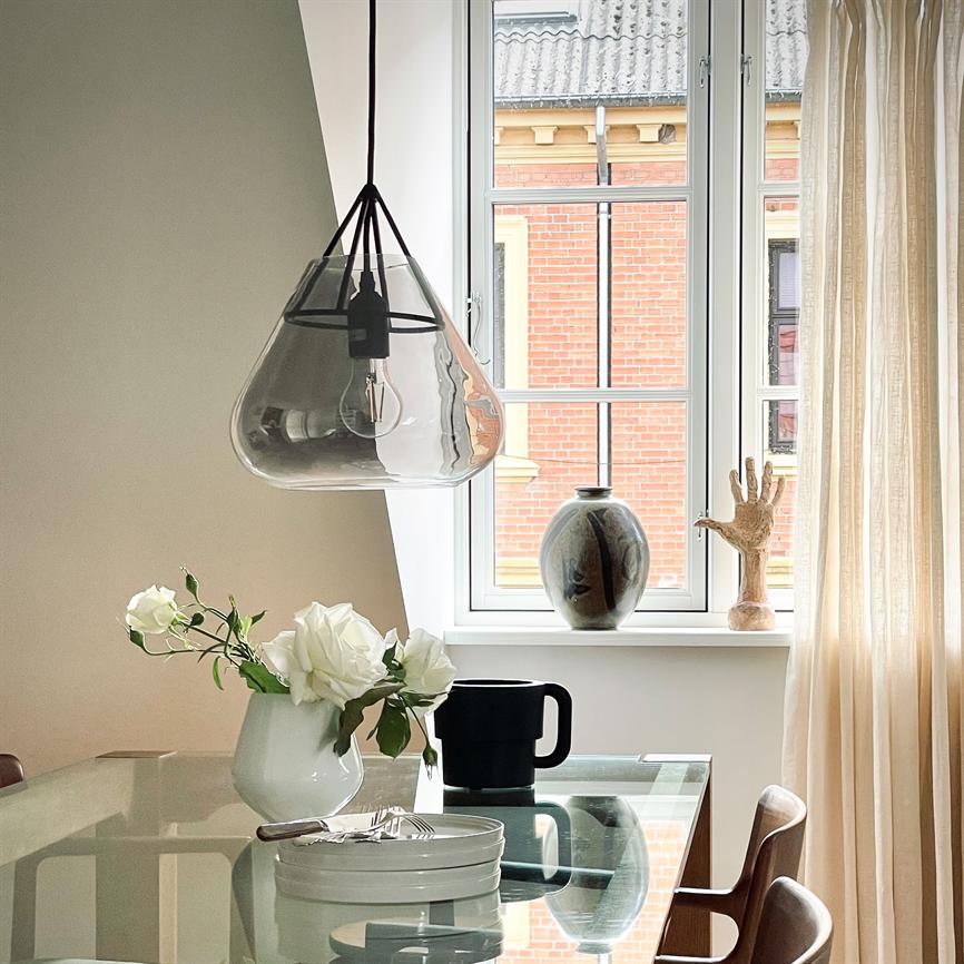 Glaspendel hænger over glasbord; på bordet en hvid vase med roser, sort krus og tallerkener. I baggrunden et vindue med murstensfacade, keramikvase og en træhånd på karmen.