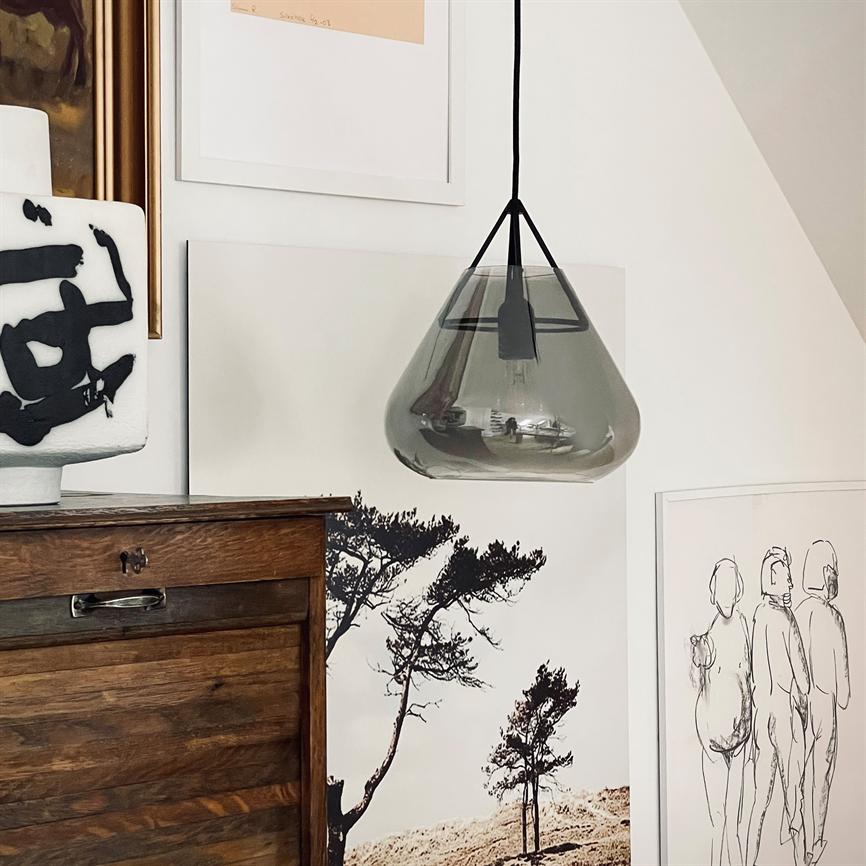 En røgfarvet glaspendel hænger over et mørkt træskab og reflekterer rummet; bagved pryder sort-hvide natur- og figurtegninger en hvid væg.