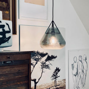 Glaspendel: hænger tændt over en mørk trækommode (handling) i en stueagtig niche, omgivet af vægkunst med et sort-hvidt træfotografi og en blækskitse af tre figurer (kontekst).