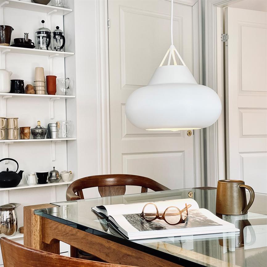 En hvid loftslampe hænger over et glasbord; på bordet ligger en åben bog med runde briller og en keramikkande; omgivet af træstole, hylde med krus og lyse døre.