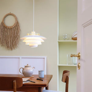 En hvid, lagdelt hængelampe lyser over et træbord i en lys spisekrog med tekande, krus, to træstole, vægdekoration af makramé og indbyggede hylder.