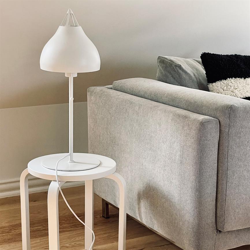 Hvid bordlampe står på et rundt hvidt sidebord; ved siden af står en lysegrå sofa med en sort-hvid, tekstureret pude i et lyst, minimalistisk stuemiljø.