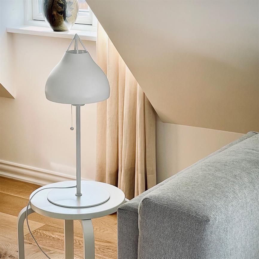 Hvid bordlampe står på et rundt sidebord, ledning slanger ned; den kaster blødt lys ved siden af en grå sofa under skrå loft, foran beige gardin og vindueskarm med vase.