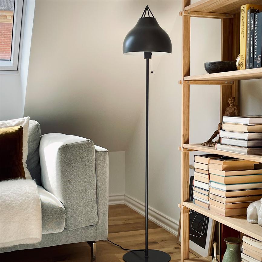 Sort gulvlampe står ved siden af en lysgrå sofa, i et solbelyst hjørne med trægulv og en åben træreol fyldt med stakke af bøger.