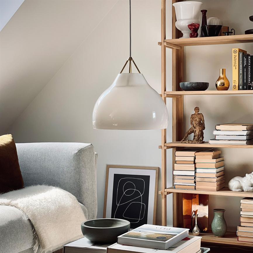 En hvid glas-hængelampe hænger over et lille sofabord; i baggrunden en lysegrå sofa med plaid og en træreol fyldt med bøger, keramik og en lille skulptur.