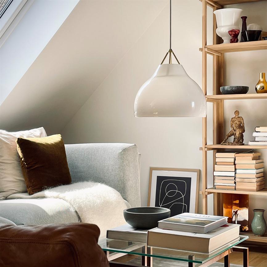 En hvid, hængende lampe lyser over et lavt sofabord; et gråt hjørnesofa med pude og tæppe står til venstre, mens en åben træreol med bøger og dekor fylder højre side.