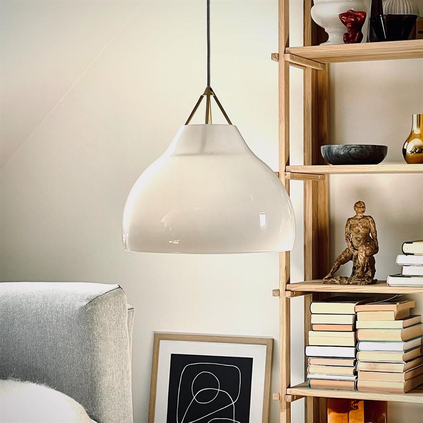 En hvid pendellampe hænger over en sofa; ved siden af står en træreol med stabler af bøger, en lille skulptur, skåle og et indrammet abstrakt print i et lyst rum.