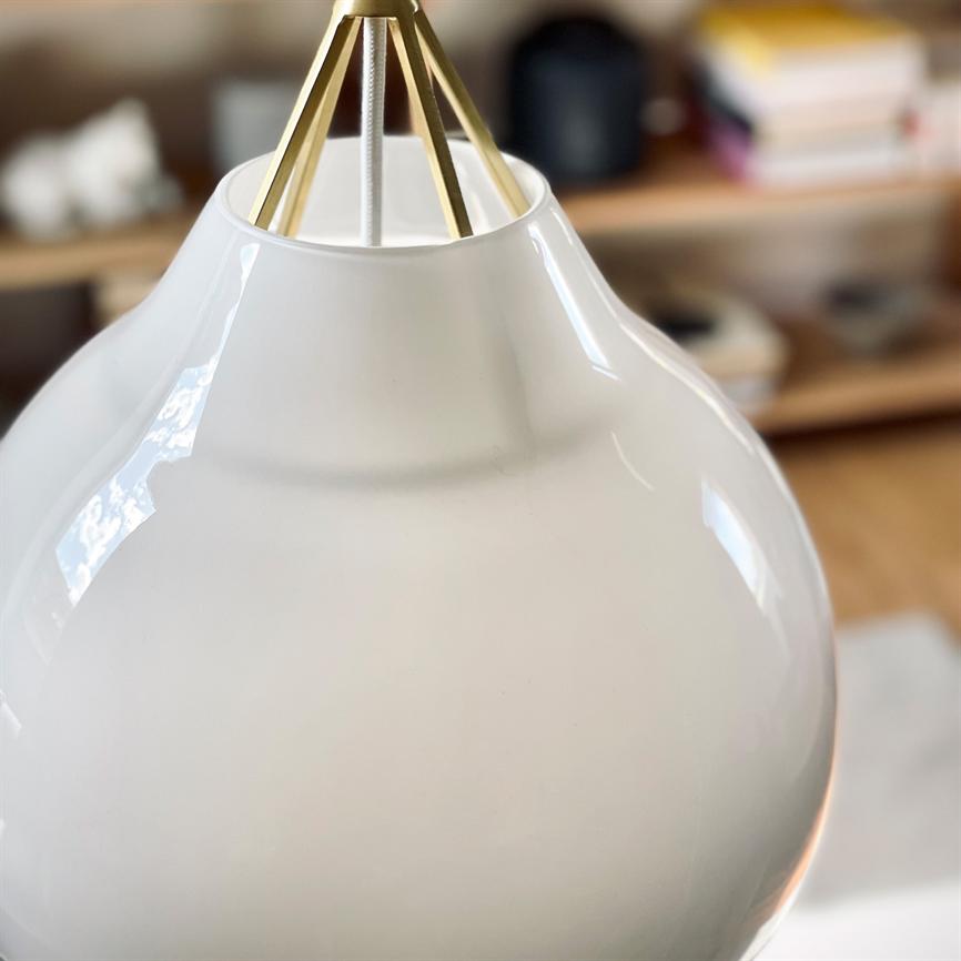 En hvid, blank pendellampe hænger i et messingophæng, reflekterer lys; i baggrunden ses sløret træreol og hylder med bøger og små genstande på et bord.