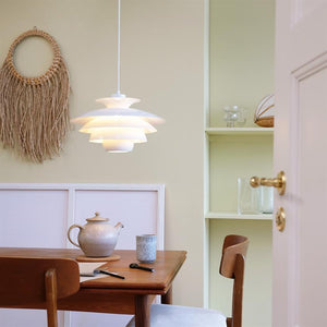 En hvid pendel lyser over et træbord med tekande og kop i et lyst spisekøkken, omgivet af et flettet vægophæng, indbyggede hylder og en åben dør med messinghåndtag.